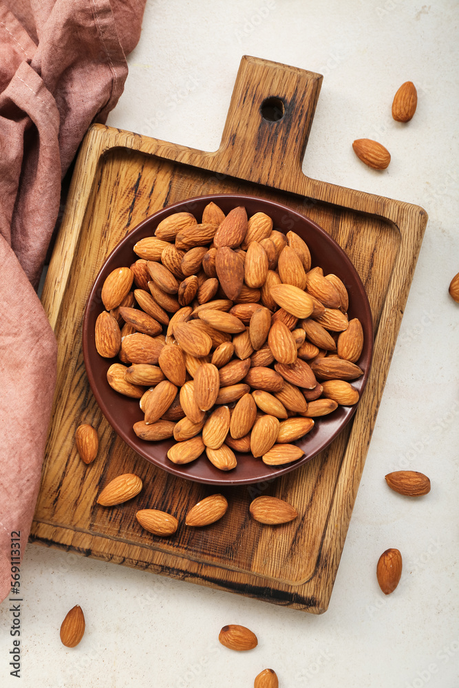 Wooden board with plate of almond nuts on light background