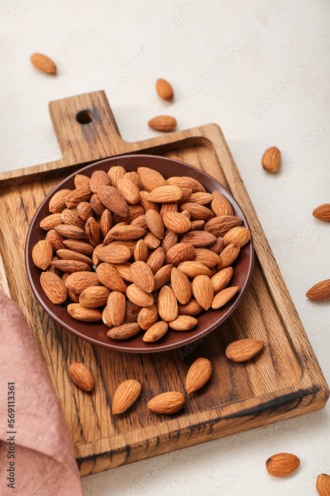 Wooden board with plate of almond nuts on light background