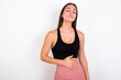 Satisfied smiling young woman wearing sportswear over white studio wall, keeps hands on belly, being in good mood after eating delicious supper, demonstrates she is full. Pleasant feeling in stomach.