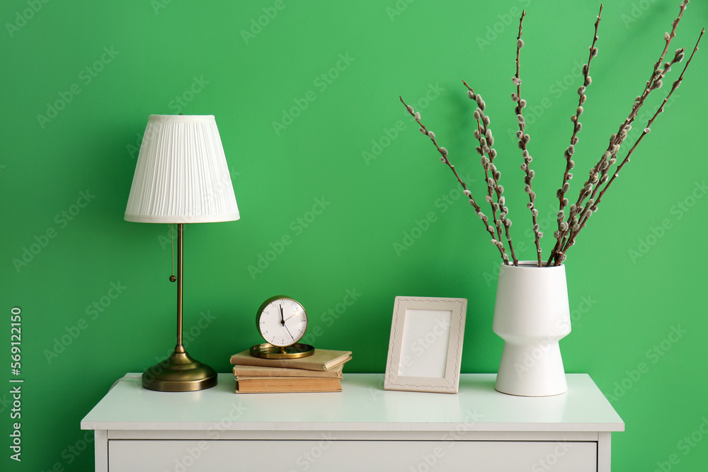 Vase with willow branches, frame, clock, books and lamp on shelf near green wall