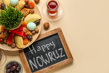 Chalkboard with text HAPPY NOWRUZ, treats, glass of tea and grass on beige background