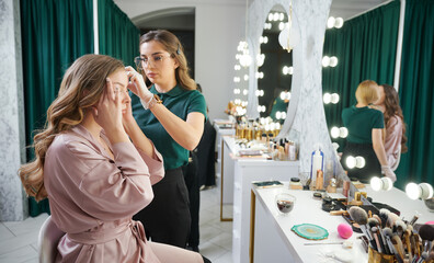 Wall Mural - Female stylist in sterile gloves holding client hair lock and pulling string while working in visage studio. Woman sitting at dressing table and touching temple while having beauty procedure in salon.