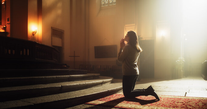 side view: christian woman getting on her knees in front of altar and starting to pray in church. de