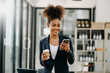 © Nuttapong punna - Confident beautiful African businesswoman typing laptop computer and digital tablet while holding coffee at office.