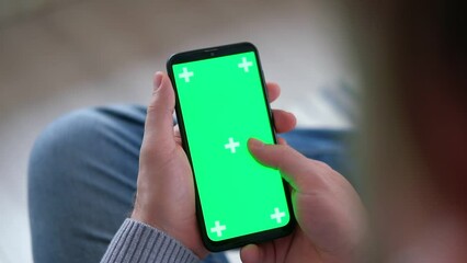 Young man sitting at home holding smartphone green mock-up screen in hand. Male person using chroma key mobile phone. Vertical mode. Touching, swiping display, tapping, surfing Internet social media