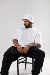 Wall Mural - Attractive african american man with a beard dressed in a white t-shirt and panama sits on a chair.