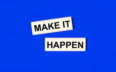 Two wooden blocks with the text MAKE IT HAPPEN on a blue background. Copy space