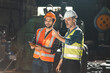 Senior male engineer training and explaining work to new employee wearing vest and safety jacket with hardhat helmet while pointing towards machine in factory and giving instructions