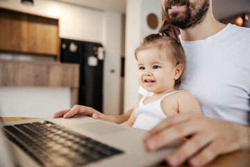 Portrait of a happy little girl sitting in her father's lap and watching while he works on a laptop.