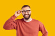 © Studio Romantic - Studio shot of happy young man in glasses. Cheerful handsome bearded male model wearing stylish eyeglasses and orange sweatshirt standing isolated on yellow background, looking at camera and smiling