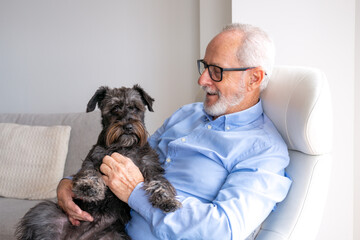 Happy Grandpa is sitting at chair in home with dog Jack Russell Terrier on his legs. Senior man 70-75 years old smiling at camera. Light window on background. Сoncept of happy old age.