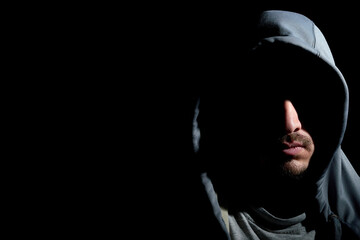 Horizontal portrait shot of mysterious young bearded hooded man with his face covered in shadows isolated on dark black background with copy space. Stranger hiding his identity, anonymous person.