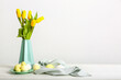 Vase with beautiful tulip flowers and plate of Easter eggs on table near light wall