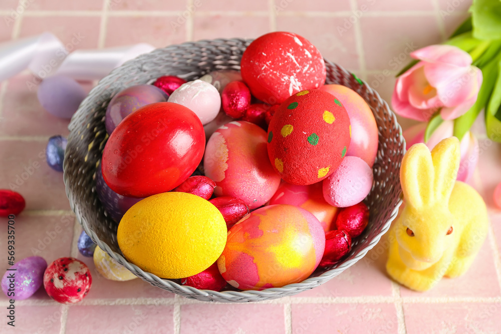 Bowl with painted Easter eggs, candies, tulips and bunny on color tile background