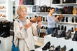 © JackF - Mature european woman who came to a shoe store demonstrates stylish loafers with heels, standing near the shelves with goods