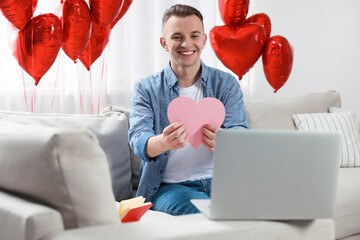 Wall Mural - Valentine's day celebration in long distance relationship. Man holding pink paper heart while having video chat with his girlfriend via laptop