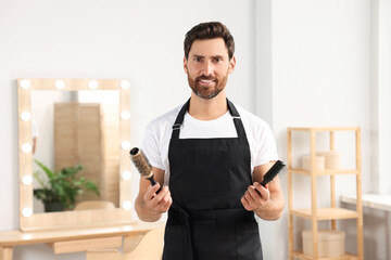 Wall Mural - Smiling hairdresser in apron holding brushes near vanity mirror in salon, space for text