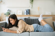 © Monster Ztudio - Happy young asian woman relax on comfortable couch at home texting messaging on smartphone, smiling girl use cellphone, browse wireless internet on gadget, shopping online from home