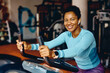 © Drazen - Happy black athletic woman cycling on stationary bike in gym and looking at camera.