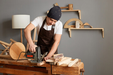 Wall Mural - Portrait of carpenter man wearing brown apron and black hat working on woodworking machines in carpentry shop, using electric jigsaw, enjoying his profession.