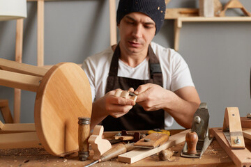 Indoor shot of handsome carpenter making a handmade wooden toy in a home workshop, enjoying wood carving, posing in his joinery, handyman wearing brown apron and black cap.