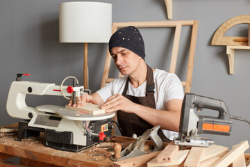 Wall Mural - Indoor shot of attractive man carpenter wearing brown apron and black cap, works with wood, using electric jigsaw for making patterns on a wooden block, working in his joinery.