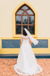 Young bride in a beautiful white wedding dress standing in front of a church window, wind blown veil