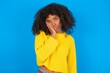 © Jihan - Sad lonely young woman with afro hairstyle wearing yellow sweater over blue wall touches cheek with hand bites lower lip and gazes with displeasure. Bad emotions