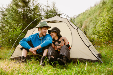 smiling tourist couple drinking tea while sitting at camping tent in forest