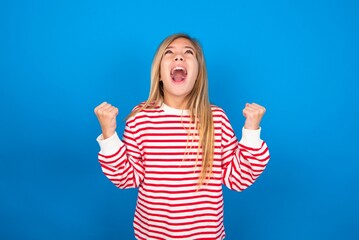 Wall Mural - caucasian teen girl wearing striped shirt over blue studio background looks with excitement up, keeps hands raised, notices something unexpected.