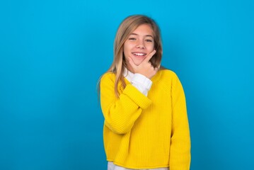 Wall Mural - caucasian teen girl wearing yellow sweater over blue studio background looking confident at the camera smiling with crossed arms and hand raised on chin. Thinking positive.