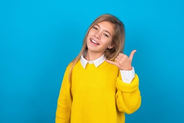 Wall Mural - caucasian teen girl wearing yellow sweater over blue studio background smiling doing phone gesture with hand and fingers like talking on the telephone. Communicating concepts.