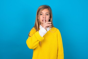 Wall Mural - caucasian teen girl wearing yellow sweater over blue studio background  makes silence gesture, keeps finger over lips. Silence and secret concept.