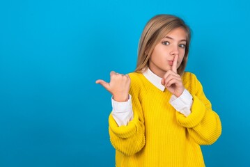 Wall Mural - caucasian teen girl wearing yellow sweater over blue studio background asking to be quiet with finger on lips pointing with hand to the side. Silence and secret concept.