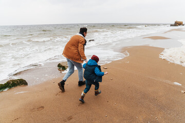 Back view of father and his son holding hands while running along beach