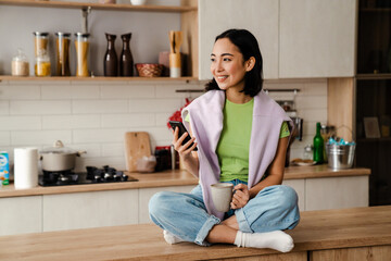 Wall Mural - Young asian woman using mobile phone and drinking coffee while sitting on table in cozy kitchen