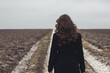 © Daria - A girl in a black coat with long curly hair walks along a dirt road.Retro vintage photo.Loneliness,thoughts and dreams.A lonely girl walks along the road.Lady in black.Photo from the back.Experiences.