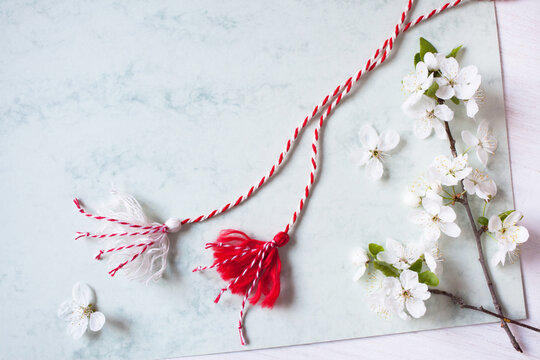 Flowering branches of cherry plum, red-white martenitsa cord with tassels on a paper, white wooden background, copy space for the holiday of March 1, Martisor, Baba Marta.