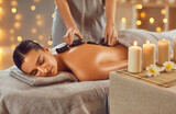 Woman enjoying exotic hot stones spa massage. Relaxed young woman lying on a spa bed while the masseuse is putting hot stones on her back. Spa treatment concept