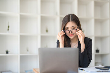 Photo of a beautiful woman holding eyeglasses while sitting at the working desk surrounded by a computer laptop, paperwork, calculator, coffee cup at office.