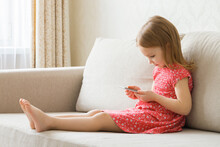 Beautiful Cute Little Girl In Red Dress Sitting On Sofa And Holding Smartphone At Home Room. Side View. Children Addiction. Spending Time Alone.