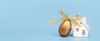 Happy Easter banner. One golden egg with a ribbon bow and white ceramic house on blue. Copy space