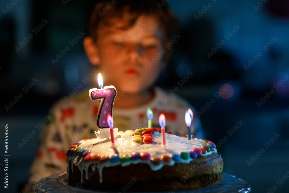 Adorable happy blond little kid boy celebrating his birthday. Child blowing seven candles on homemade baked cake, indoor. Birthday party for school children, family celebration of 7 years