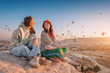 © EdNurg - Girl friends sitting on a clifftop viewpoint and admiring view of majestic flying hot air balloons in Cappadocia
