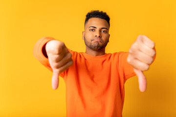 Unhappy young Indian man showing rejection and negative with thumbs down gesture isolated on yellow. Sad guy demonstrates disagreeing, bad review, dislike gesture