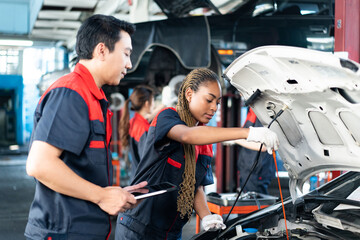 Selective focus of a young African female mechanic in uniform, checking car engine oil level with open hood, with a senior Asian male mechanic holding a tablet standing beside her in a blurred garage.