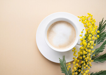 Wall Mural - A spring bouquet with yellow mimosa flowers and a cup of coffee  cappuccino on a beige background. Concept of 8 March, happy women's day.