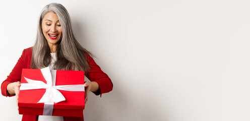 Wall Mural - Happy elderly woman with grey hair, receive present, looking at red gift box and smiling surprised, standing over white background