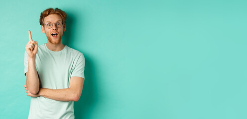 Wall Mural - Excited young man with ginger hair in glasses, raising index finger, pitching an idea, standing over mint background