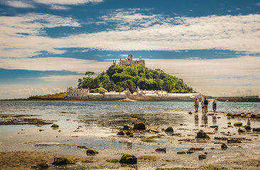 Group of people looking at tidal island in Cornwall, England, while standing at the edge of coming tide water on sunny summer day; rocks on foreground; blue sky with clouds in background